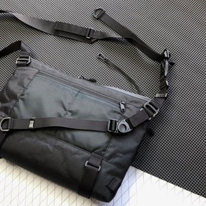 May include: A black crossbody bag with a zippered pocket on the front and adjustable straps. The bag is made of a durable, water-resistant material and has a sleek, modern design. Shoulder Bag, Messenger Bag, Techwear, X Pac, Sling Bag, EDC Bag, Commuter bag, lightweight bag, Cycling Bag, Laptop Bag, MacBook bag, D4bgs