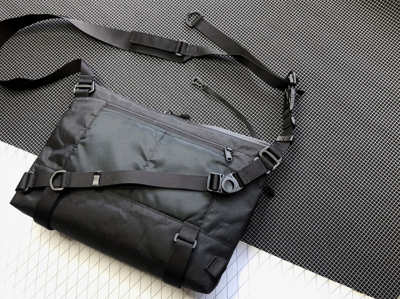 May include: A black crossbody bag with a zippered pocket on the front and adjustable straps. The bag is made of a durable, water-resistant material and has a sleek, modern design. Shoulder Bag, Messenger Bag, Techwear, X Pac, Sling Bag, EDC Bag, Commuter bag, lightweight bag, Cycling Bag, Laptop Bag, MacBook bag, D4bgs