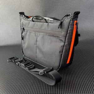 May include: A gray messenger bag with a black adjustable strap. The bag has a zippered main compartment and a front zippered pocket. An orange stripe runs down the side of the bag. The bag is made of a durable, lightweight material.