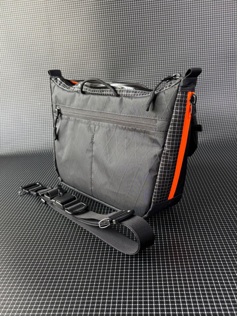 May include: A gray messenger bag with a black adjustable strap. The bag has a zippered main compartment and a front zippered pocket. An orange stripe runs down the side of the bag. The bag is made of a durable, lightweight material.