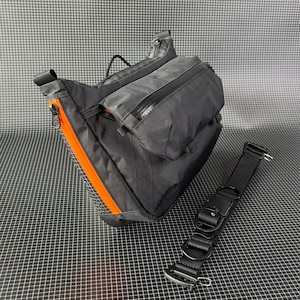 May include: Black crossbody bag with orange zipper accents and a detachable black strap. The bag features multiple zippered compartments and a durable, textured material. The bag is designed for carrying essentials.