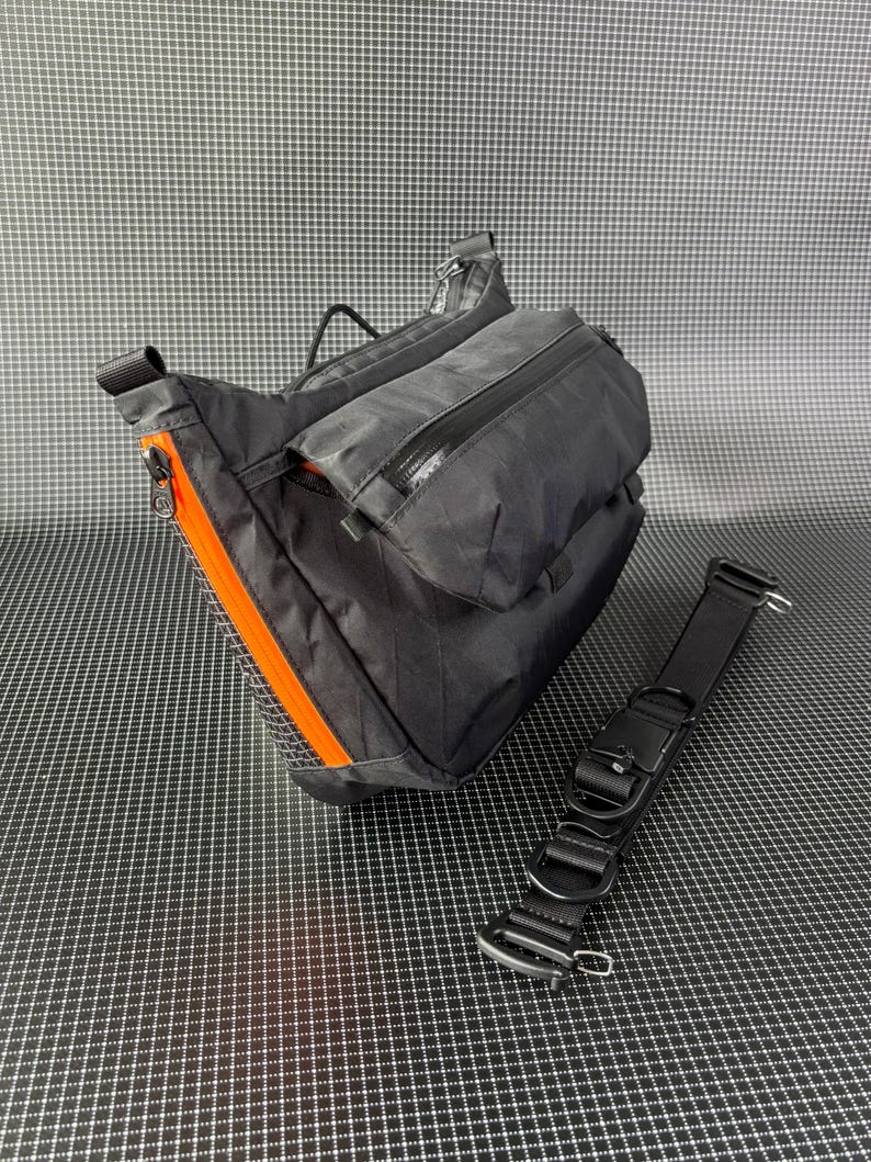 May include: Black crossbody bag with orange zipper accents and a detachable black strap. The bag features multiple zippered compartments and a durable, textured material. The bag is designed for carrying essentials.