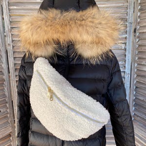 May include: A black puffer jacket with a fur-trimmed hood and a white, fluffy bum bag with a gold zip. The bum bag is made of a soft, textured material and is worn across the body.