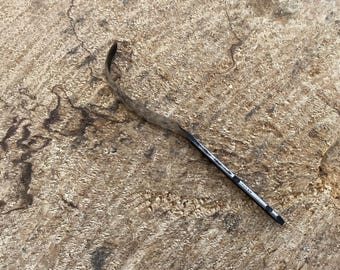 May include: A black and silver pen lies on a textured, light brown surface. The pen has a black barrel with silver accents and a black curved tip. The surface has dark brown markings and a rough, natural appearance.