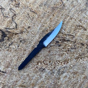May include: A small, hand-forged knife with a dark handle and a light blue blade. The knife is set against a textured, light brown wooden surface, showcasing the craftsmanship of the blade.