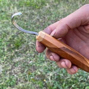 May include: A wood carving hook knife with a curved blade. The handle is made of light brown wood with a faceted design. The metal blade curves upward, creating a hook shape. The knife is designed for detailed wood carving.