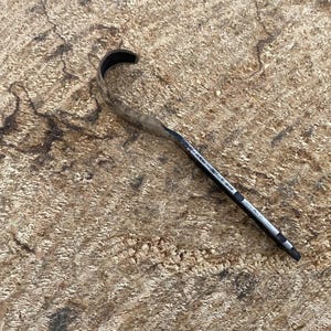Könnte beinhalten: Ein schwarzes und silbernes Metallwerkzeug mit gebogenem Ende liegt auf einer strukturierten, hellbraunen Oberfläche. Das Werkzeug hat einen schlanken Körper und eine spitze Spitze. Der Hintergrund ist ein raues, hellbraunes Material mit dunkelbraunen Markierungen.