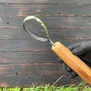 Puede incluir: Una herramienta con mango de madera y hoja metálica curvada. El mango es de color marrón claro y la hoja tiene forma de lazo. La herramienta se sostiene sobre un fondo de madera oscura.