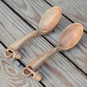 Puede incluir: Dos cucharas de madera de color marrón claro, con mangos largos y cuencos ovalados. Las cucharas parecen talladas a mano, cada una con un lazo en el extremo del mango. Se muestran sobre una superficie de madera.