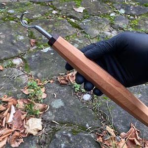 Könnte beinhalten: Ein Holzschnitzhakenwerkzeug mit einer gebogenen Metallklinge und einem facettierten Holzgriff. Der Griff ist hellbraun mit einem dunkelbraunen Akzent in der Nähe der Klinge. Das Werkzeug wird vor einem Hintergrund aus Stein und Herbstlaub gehalten.
