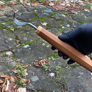 Könnte beinhalten: Ein Holzschnitzmesser mit gebogener Klinge. Der Griff ist hellbraun und hat ein facettiertes Design. Die silberne Klinge ist gebogen. Das Werkzeug wird von einer Person mit schwarzem Handschuh gehalten.