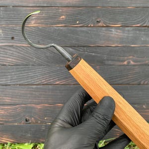 Könnte beinhalten: Ein Holzschnitzhakenmesser mit gebogener Klinge. Der Griff ist aus hellem Holz und hat eine dunkle Metallklinge. Das Messer wird von einer behandschuhten Hand vor einem dunklen Holzhintergrund gehalten.