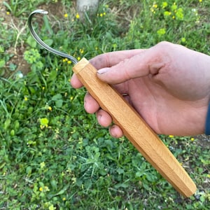 Könnte beinhalten: Ein Schnitzhaken-Werkzeug mit einem Holzgriff und einer gebogenen Metallklinge. Der Griff ist hellbraun und hat ein facettiertes Design. Das Werkzeug wird in einer Hand gehalten.