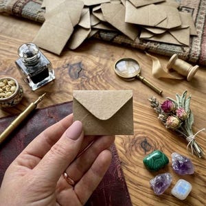 May include: A small, rectangular, light brown kraft paper envelope held in the center. The scene includes a glass inkwell, a gold-toned fountain pen, a magnifying glass, a small bouquet of dried flowers, and four polished gemstones.