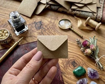May include: A small, rectangular, light brown kraft paper envelope held in the center. The scene includes a glass inkwell, a gold-toned fountain pen, a magnifying glass, a small bouquet of dried flowers, and four polished gemstones.
