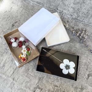 Pode incluir: Um alfinete de lapela prateado com uma flor branca de cinco pétalas e centro escuro, apresentado numa caixa preta. Ao lado, uma caixa castanha contém um grupo de pequenos pendentes decorativos florais e de bagas em vermelho, branco e verde.