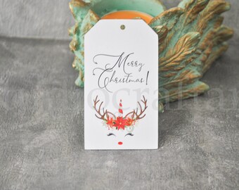 Pode incluir: Etiqueta de presente branca com uma ilustração de rena unicórnio. A rena tem chifres vermelhos e brancos, um nariz vermelho e um chifre de pirulito vermelho e branco. A etiqueta diz "Feliz Natal!" em escrita cursiva preta.