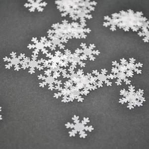以下が含まれることがあります： 暗い灰色の表面に散らばった白い雪の結晶の紙吹雪。雪の結晶は薄くて半透明な素材でできており、さまざまなサイズです。