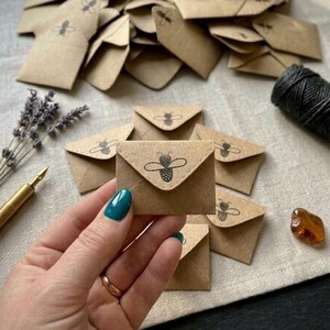 May include: A collection of small, square, light brown kraft paper envelopes, each featuring a black ink stamp of a bee on the triangular flap. One envelope is held in the foreground, while others are scattered behind it on a textured fabric.