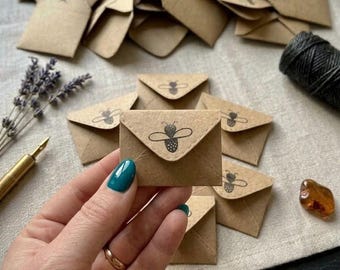 May include: A collection of small, square, light brown kraft paper envelopes, each featuring a black ink stamp of a bee on the triangular flap. One envelope is held in the foreground, while others are scattered behind it on a textured fabric.