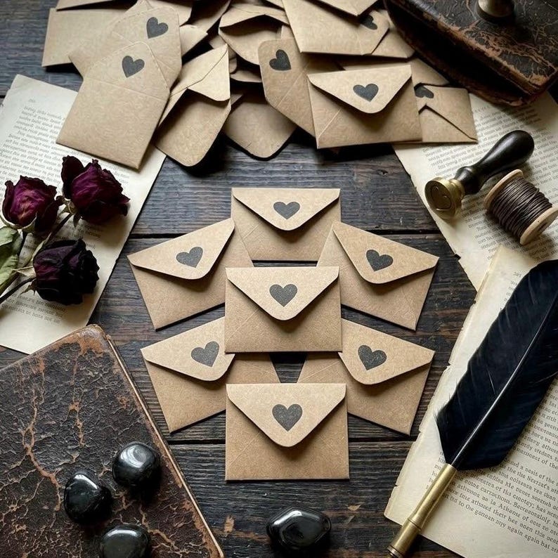 May include: A collection of small brown kraft paper envelopes, each featuring a black heart icon on the flap. The envelopes are arranged on a dark wooden surface alongside dried dark red roses, a black quill pen, a wax seal stamp, and a vintage book.