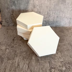 Pode incluir: Uma pilha de caixas hexagonais brancas. As caixas são feitas de um material fosco e são dispostas sobre uma superfície cinza texturizada. As caixas estão vazias e prontas para uso.