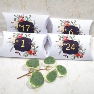 Könnte beinhalten: Vier weiße Kissenboxen mit den goldenen Zahlen 1, 6, 17 und 24, für einen Adventskalender. Jede Box hat eine dunkelblaue Form mit einem floralen Design aus roten Beeren, grünen Blättern und weißen Blumen. Ein Zweig mit grünen Blättern ist im Vordergrund.
