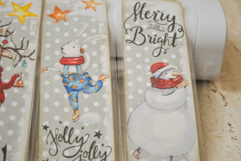 May include: Four Christmas-themed bookmarks with a gray background and white polka dots. The bookmarks feature a reindeer, a rabbit ice skating, a snowman, and the text "Merry and Bright".