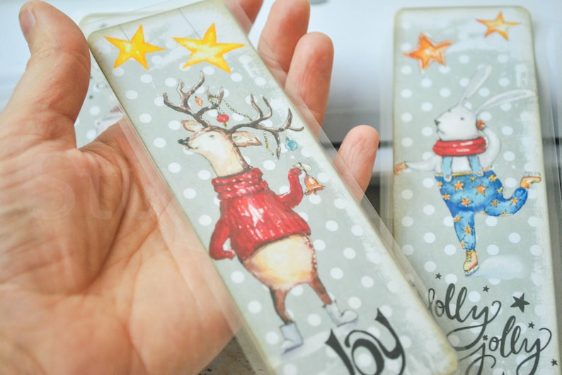 May include: Two Christmas-themed bookmarks. The first bookmark features a cartoon deer wearing a red sweater and holding bells. The second bookmark features a cartoon bunny wearing blue overalls and ice skates. Both bookmarks have a gray background with white polka dots and the words "joy" and "jolly" printed on them.