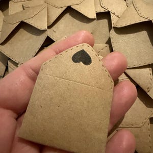 May include: A small, light brown kraft paper envelope featuring a pointed flap with a black heart icon stamped on the center. The envelope has visible stitched detailing along the edges and is part of a larger collection of identical envelopes.