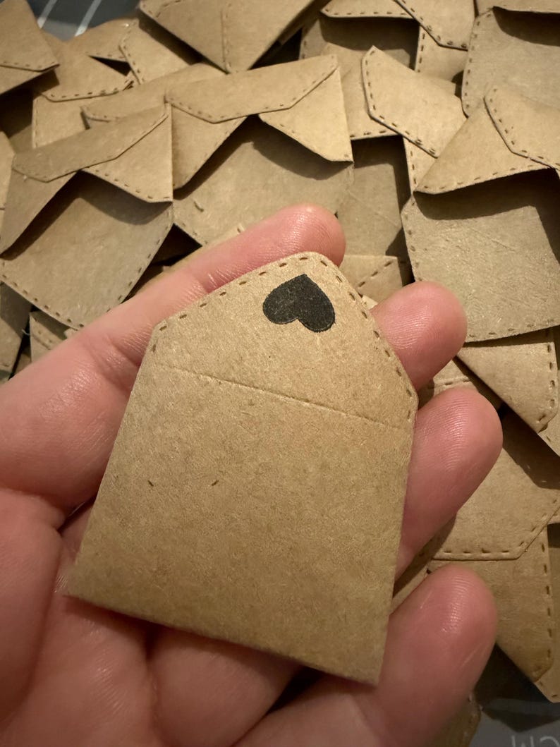 May include: A small, light brown kraft paper envelope featuring a pointed flap with a black heart icon stamped on the center. The envelope has visible stitched detailing along the edges and is part of a larger collection of identical envelopes.