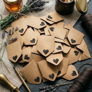 May include: A collection of small, light brown paper envelopes, each featuring a stamped black heart design. The envelopes are arranged in a pile alongside a metal dip pen, a bundle of dried lavender, and a spool of dark brown twine.
