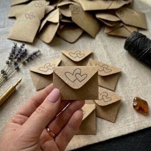 Op de afbeelding: Een verzameling kleine, vierkante enveloppen van lichtbruin kraftpapier, elk voorzien van een gestempelde zwarte omtrek van twee in elkaar grijpende harten op de klep. De enveloppen liggen op een stapel, één wordt vooraan vastgehouden.