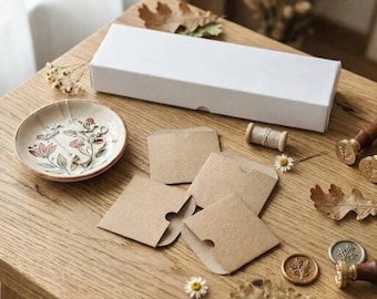 May include: A white rectangular box, a small ceramic dish with a floral pattern containing a silver necklace, several brown paper envelopes, a spool of thread, two metal wax seals, and dried oak leaves arranged on a wooden surface.