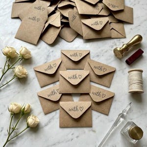 May include: A collection of brown kraft paper envelopes, each stamped with the text "with ♡". The envelopes are arranged alongside cream-colored roses, a gold-toned wax seal stamp, a spool of white twine, and a glass dip pen with an ink bottle.