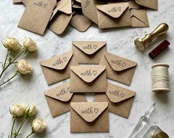 May include: A collection of brown kraft paper envelopes, each stamped with the text "with ♡". The envelopes are arranged alongside cream-colored roses, a gold-toned wax seal stamp, a spool of white twine, and a glass dip pen with an ink bottle.