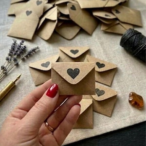 May include: A collection of small, light brown kraft paper envelopes, each featuring a black heart icon printed on the triangular flap. The envelopes are arranged in a scattered pile, with one envelope held in the foreground.