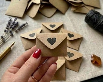 May include: A collection of small, light brown kraft paper envelopes, each featuring a black heart icon printed on the triangular flap. The envelopes are arranged in a scattered pile, with one envelope held in the foreground.
