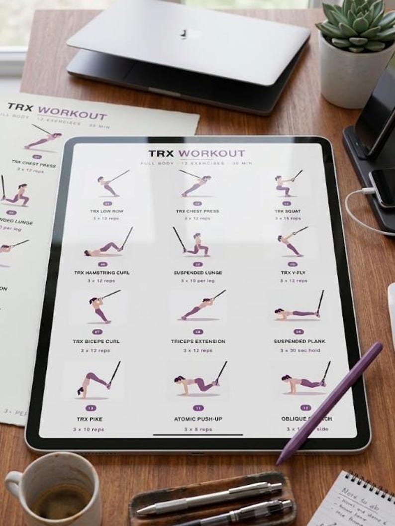 May include: A digital tablet displays a TRX workout routine with illustrations of exercises. The tablet is on a wooden surface, accompanied by printed workout sheets, a laptop, a pen, and a coffee mug. A small succulent plant is also visible.