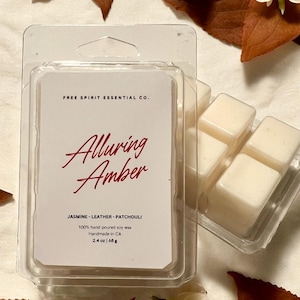 May include: A white wax melt in a clear plastic container. The wax melt is divided into four squares. The container has a label that reads "FREE SPIRIT ESSENTIAL CO. Alluring Amber JASMINE-LEATHER-PATCHOULI 100% hand-poured soy wax Handmade in CA 2.4 oz | 68 g"