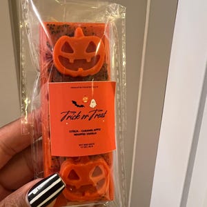 May include: A Halloween-themed soy wax melt package. The orange wax melts are shaped like jack-o'-lanterns. The package has a label that reads "Trick or Treat" with scent notes of citrus, caramel apple, and whipped vanilla.