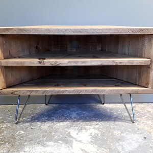 K&ouml;nnte beinhalten: Ein Eck-TV-St&auml;nder aus recyceltem Holz mit zwei Regalen und Haarnadelbeinen. Der St&auml;nder ist hellbraun und hat einen rustikalen Look.