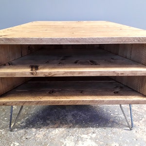 K&ouml;nnte beinhalten: Ein Eck-TV-St&auml;nder aus recyceltem Holz mit zwei Regalen und Haarnadelbeinen. Der St&auml;nder ist in einem hellen Braunton gebeizt.