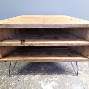 K&ouml;nnte beinhalten: Ein Eck-TV-St&auml;nder aus recyceltem Holz mit zwei Regalen und Haarnadelbeinen. Der St&auml;nder ist hellbraun und hat einen rustikalen Look.