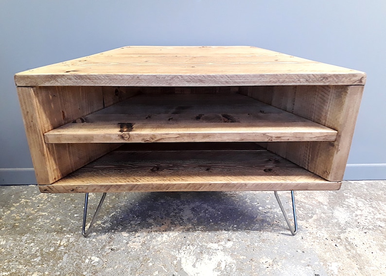 K&ouml;nnte beinhalten: Ein Eck-TV-St&auml;nder aus recyceltem Holz mit zwei Regalen und Haarnadelbeinen. Der St&auml;nder ist hellbraun und hat einen rustikalen Look.