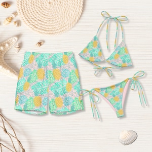 May include: A matching swimwear set comprising shorts and a bikini. The set features a repeating pattern of yellow pineapples, green leaves, and blue accents on a light pink background. The bikini top and bottom have tie fastenings.