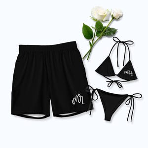 May include: Black swim trunks with the text mr. in white script on the lower leg, paired with a black bikini set featuring the text mrs. in white script on the triangle top, accompanied by a stem of two white roses with green leaves.