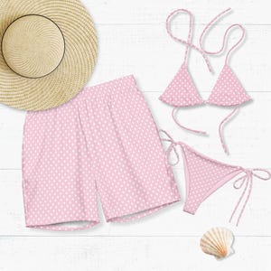 Könnte beinhalten: Ein hellrosa Bikini-Set mit weißen Punkten, passende Badeshorts und ein Sonnenhut aus Stroh. Eine Muschel ist ebenfalls vorhanden. Die Artikel sind auf einer weißen Holzoberfläche angeordnet, was ein Strand- oder Urlaubsthema suggeriert.