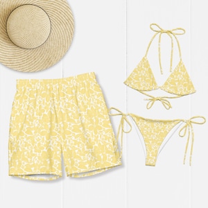 Könnte beinhalten: Ein Strohhut, eine Badehose und ein zweiteiliger Bikini, alle mit einem passenden gelb-weißen Blumenmuster. Das Bikinioberteil hat dreieckige Cups mit Bindebändern, und das Bikiniunterteil verfügt über seitliche Bindebänder.