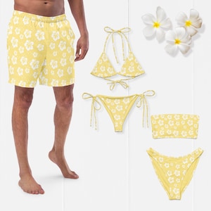 Op de afbeelding: Gele zwemkleding set en shorts met witte bloempatronen. De set bevat een bikinitop, twee bikinibroekjes en een bandeau top. De shorts worden door een persoon gedragen. Witte bloemen op de achtergrond.